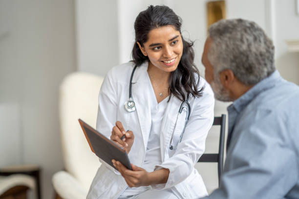 A young female doctor of Middle Eastern decent, sits with a senior patient in the comfort of his own home during a Homecare visit.  She is wearing a white lab coat, has a stethoscope around her neck and is holding out a tablet as she shows the gentleman his recent test results.   The senior is leaning in to look at the screen as he listens attentively.